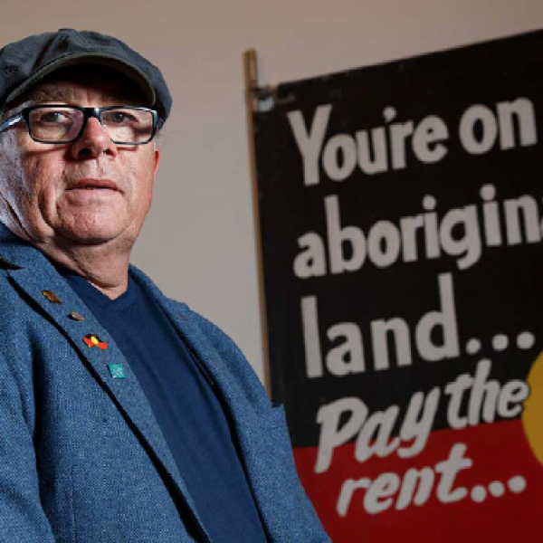 Prof Maynard standing side on looking at the camera unsmiling with a sign to his left saying, 