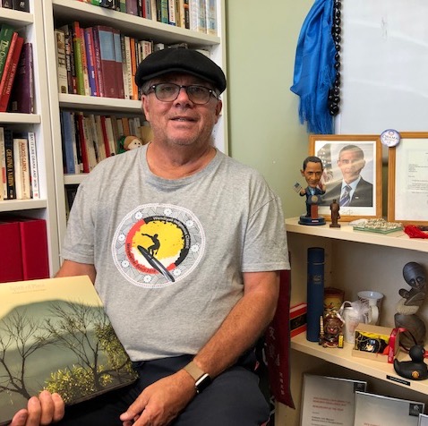Professor Maynard sitting holding a book in front of a cabinet of memorabilia collected as an historian
