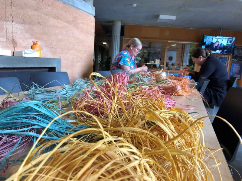 Different coloured raffia in the foreground. In the background 2 women weaving