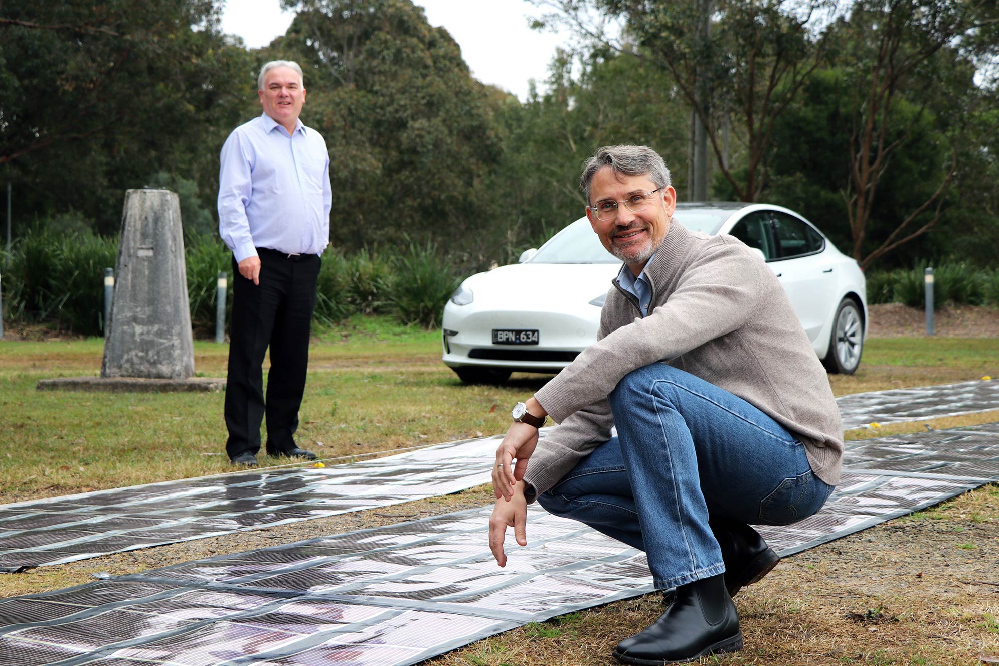 Paul Dastoor is crouched in foreground with printed solar laid on the ground and Stuart McBain in the background with an electric vehicle