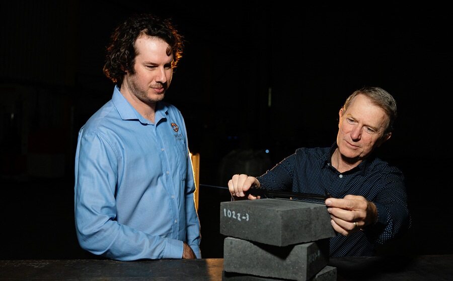 A man on the left is wearing a blue shirt. The man on the right is measuring two stacked bricks. The background is black