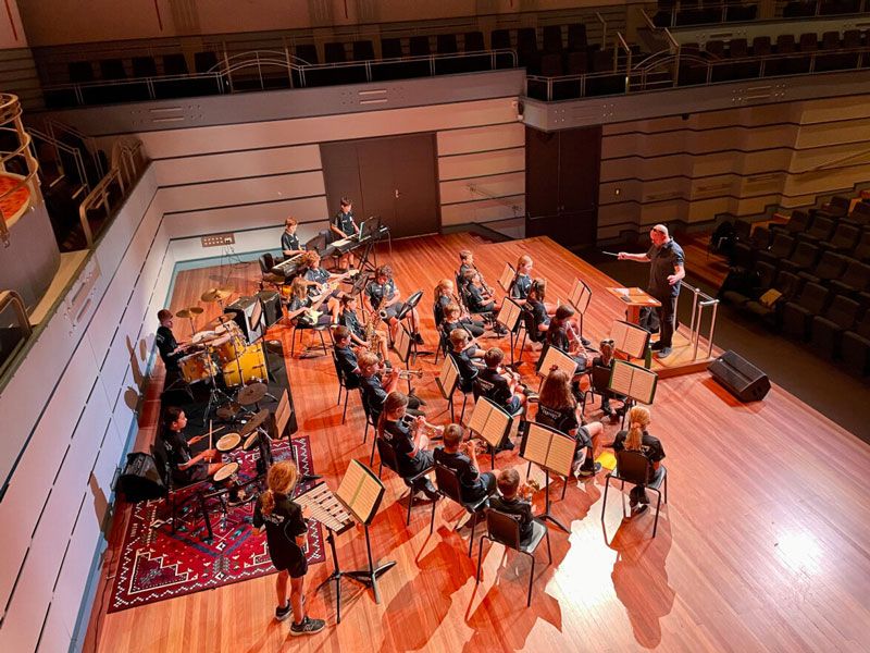 A school class of students playing in a bad with a conductor at the Conservatorium music hall