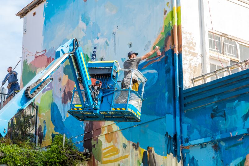Colourful mural being painted on the side of a building by a man on a crane and man on street level