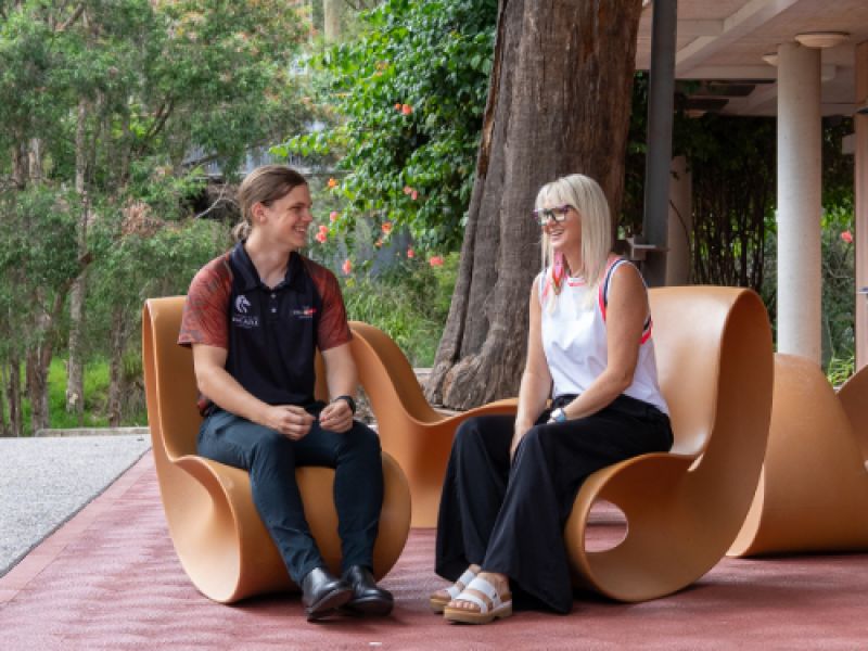 A woman and a young man sit facing each other on curved chairs outside, with greenery around them.