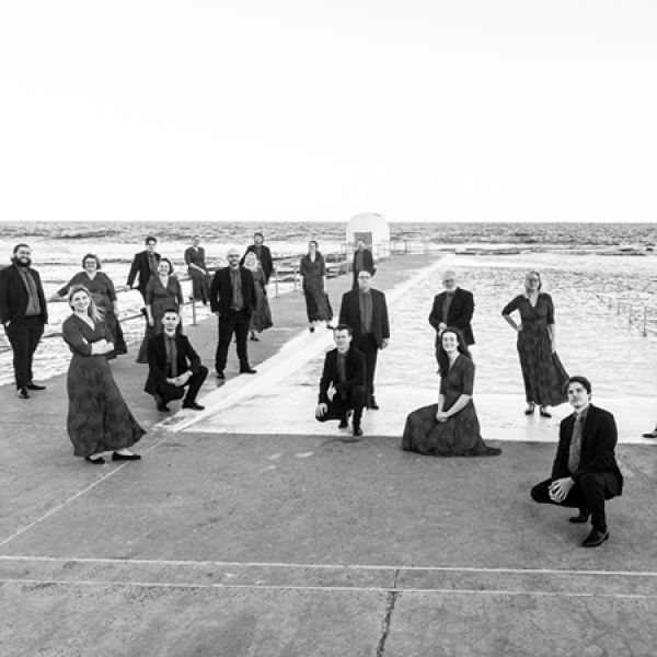A group of formally dressed performers poses on a seaside promenade in a black-and-white photo, with the ocean and horizon stretching behind them.