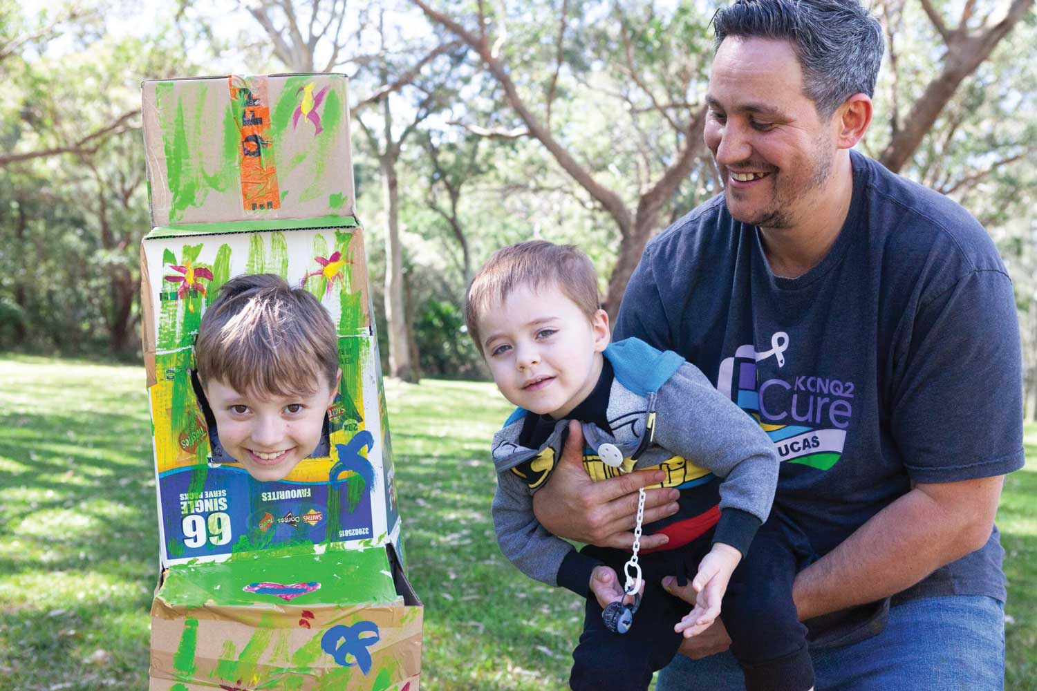 SNUG retreats, such as the one shown here, celebrate all family members. Music and art therapy, outdoor fun and sensory play are always a big hit with the kids while parents enjoy connecting with other families in the ‘same boat’. A smiling child's head pokes through a painted cardboard box, with another child and an adult male next to them smiling