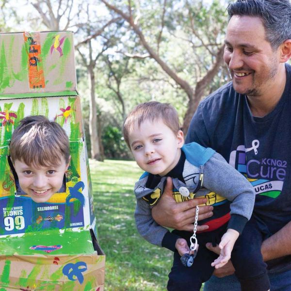 A smiling child's head pokes through a painted cardboard box, with another child and an adult male next to them smiling. Snug celebrates 15 years.