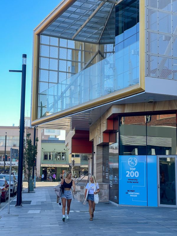 Two women with surfboards walking down a street surrounded by buildings