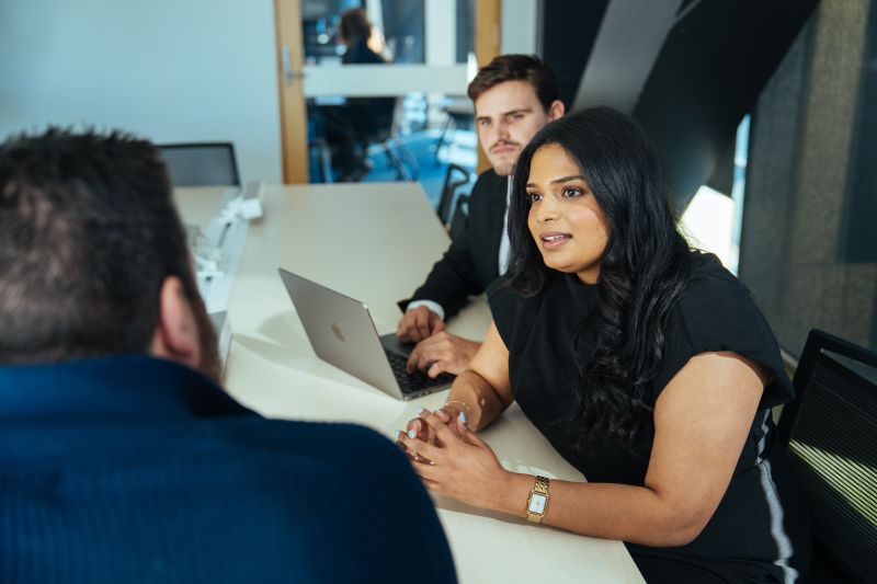 Three people sitting around a desk in an office,  Two people sit on one side, facing a third person across the table. 
