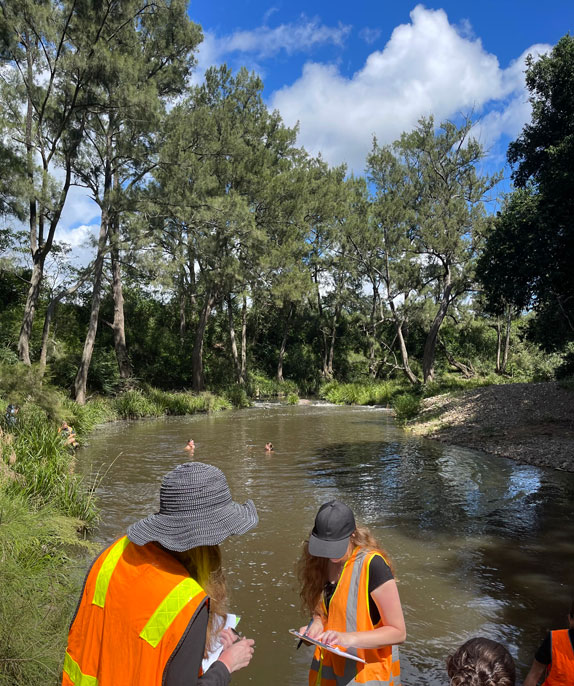 Two students stand in the foreground with black hats, hi-vis vests, and clipboards. Two students can be seen in the water in the background.