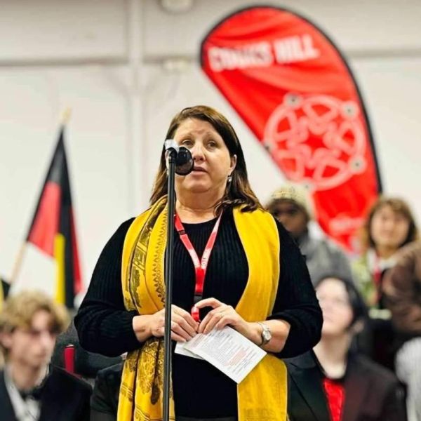 A woman in a yellow scarf speaking at a microphone. NAIDOC 2025 The Next Generation: Strength, Vision & Legacy