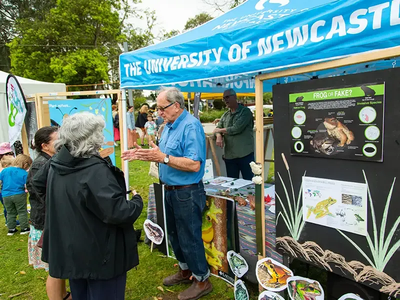 A University representative talks to festival goers in front of a University festival stall featuring infortmational posters about frogs.