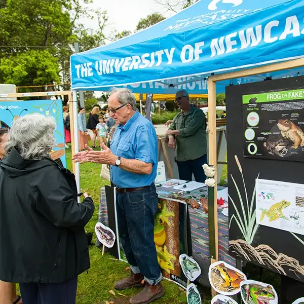 A University representative talks to festival goers in front of a University festival stall featuring infortmational posters about frogs.
