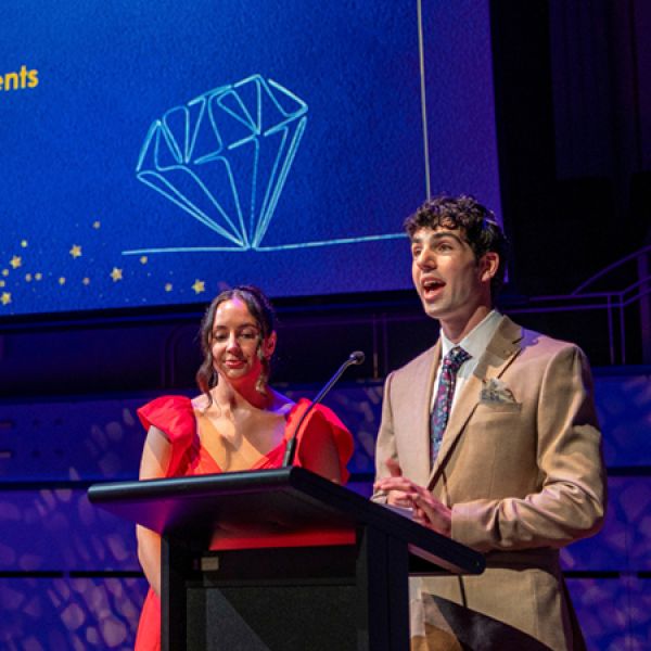 Two people stand at a lectern on a brightly lit stage, with a large blue screen displaying a diamond illustration behind them.