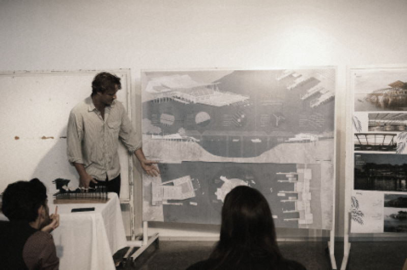 Mitchell Hosking presents architectural plans on a board to a seated audience. The atmosphere is focused, with detailed drawings and diagrams on display.