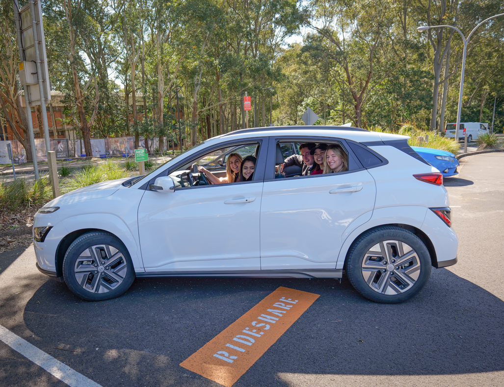 Students sitting in a white car that is parked in a designated rideshare spot in a parking area surrounded by tall trees