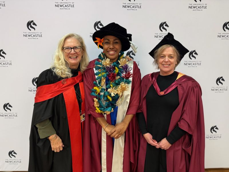 Three women in front of a university logo backdrop wearing university gowns