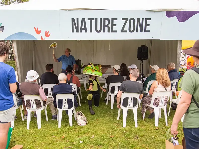 A University representative holding a cardboard cutout of a frog giving a presentation in a festival stall labelled 'Nature Zone'