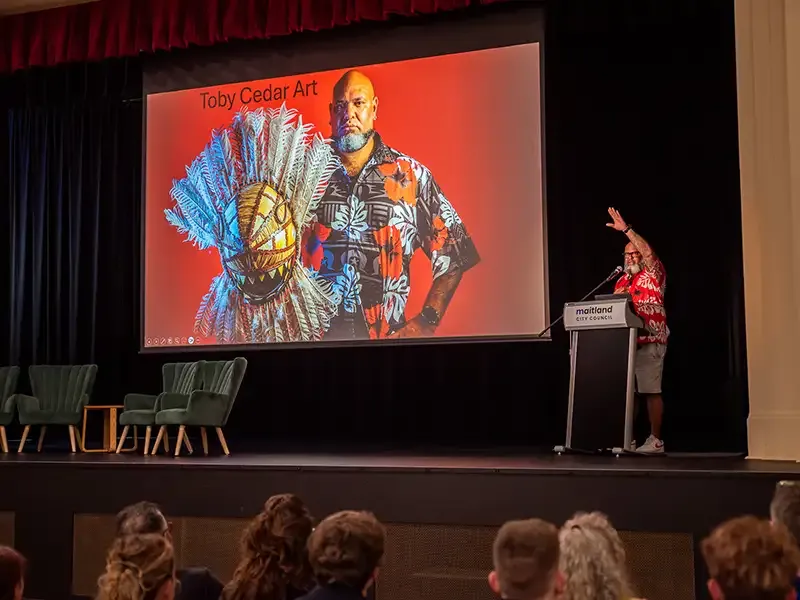 A presenter on a stage next to a slide labelled 'Toby Cedar Art'