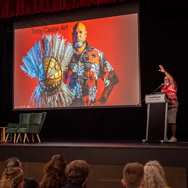 A presenter on a stage next to a slide labelled 'Toby Cedar Art'