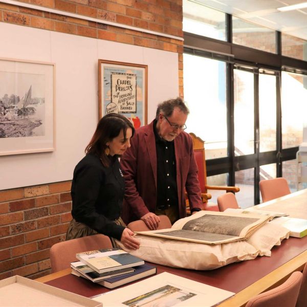 Candid picture of Dr Carl Caulfield and Paige Wright viewing a black and white, hard copy archival image in a room within the University's Library. In the background of the image there is a window, bricks, a bookcase and two out of focus picture frames.. New creative in residence inspired by history.