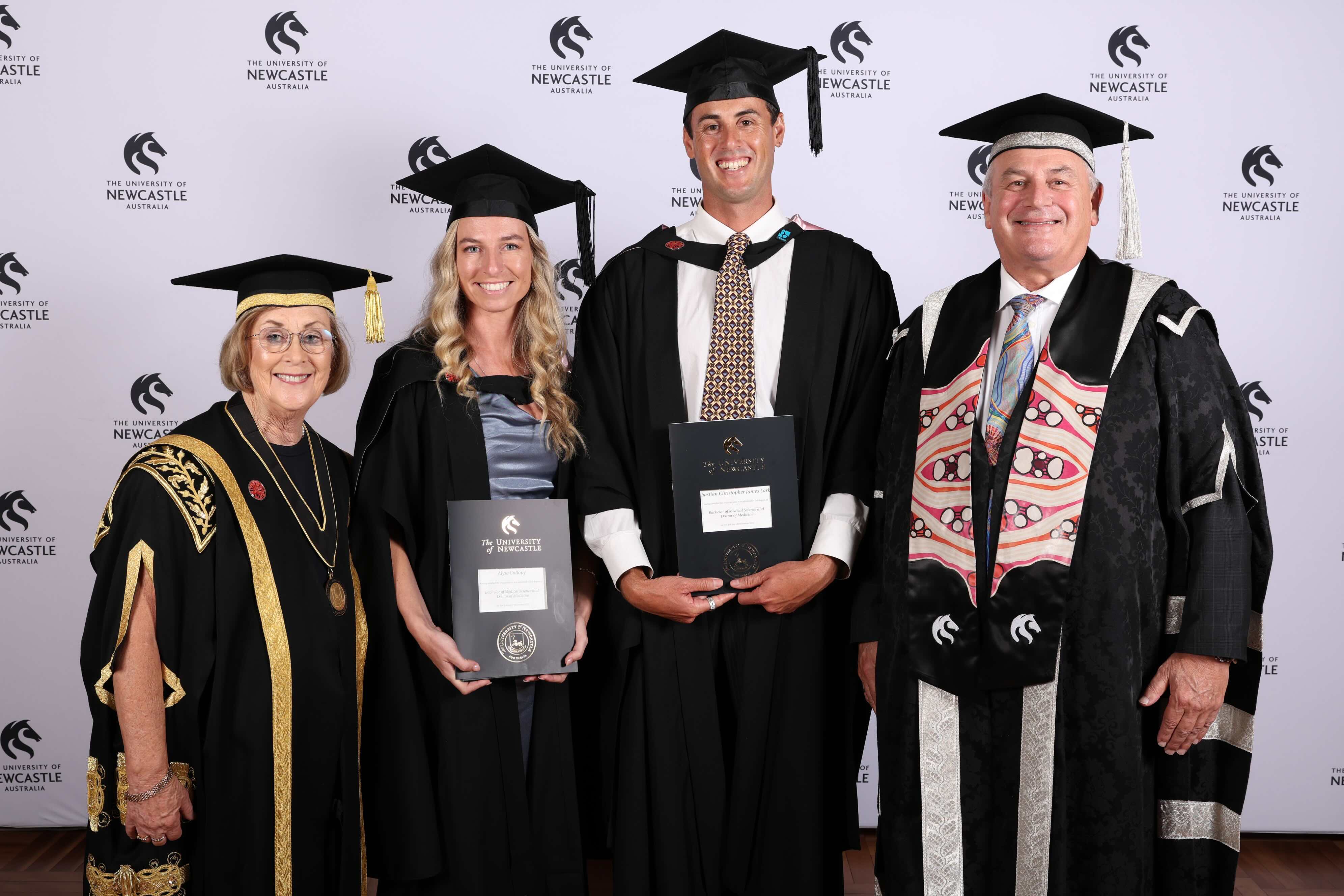 Four people with caps and gown stand looking at the camera. Two graduates are in the centre holding their degrees
