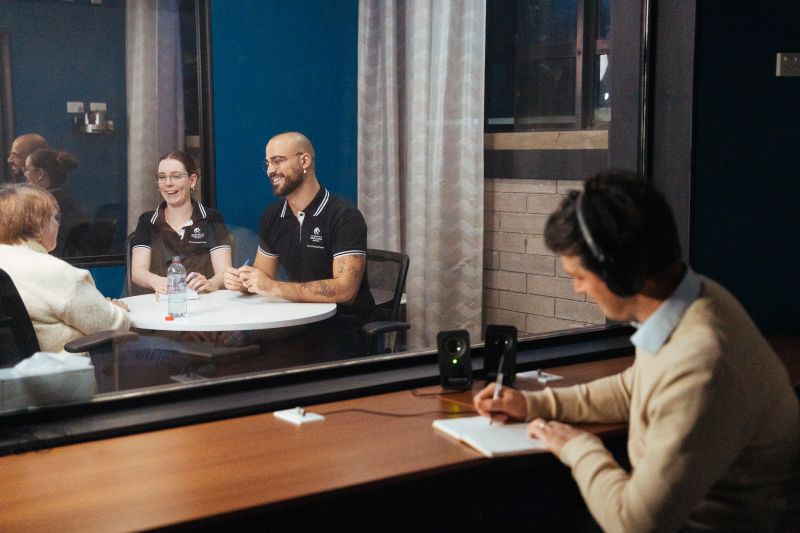 A group of three people seated around a table, engaged in a conversation. Behind a glass partition, another person wearing headphones observes and takes notes.