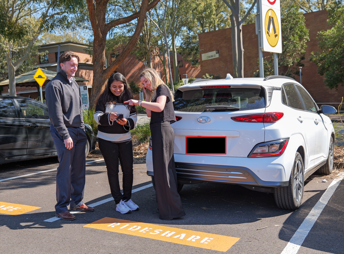 Three students standing behind a white, parked rideshare car while looking at a smartphone