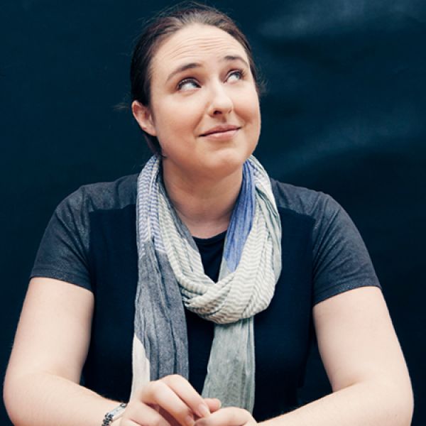 A person sits against a dark backdrop wearing a short‑sleeved top and a light, patterned scarf, with their hands resting in front of them.