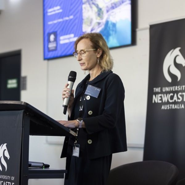 Woman with blonde hair, wearing glasses, standing behind a lectern and holding a microphone. Hunter Insights Series explores the regional impacts of NDIS reform