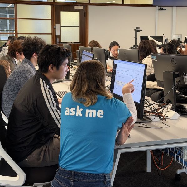 An ASKUON helper with a blue 'ask me' shirt sits at a computer with a commencing student. 