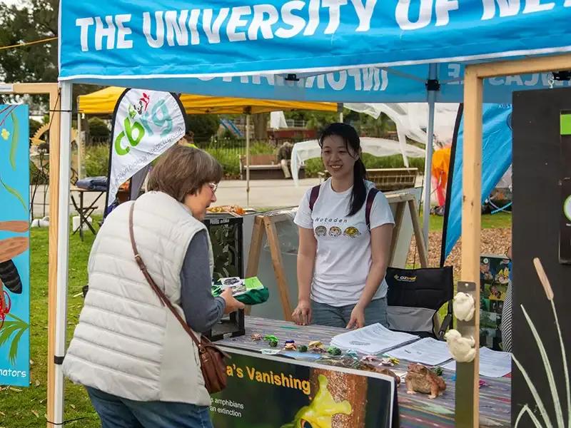 A University representative behind a counter at a University festival stall talking to a festival attendee.