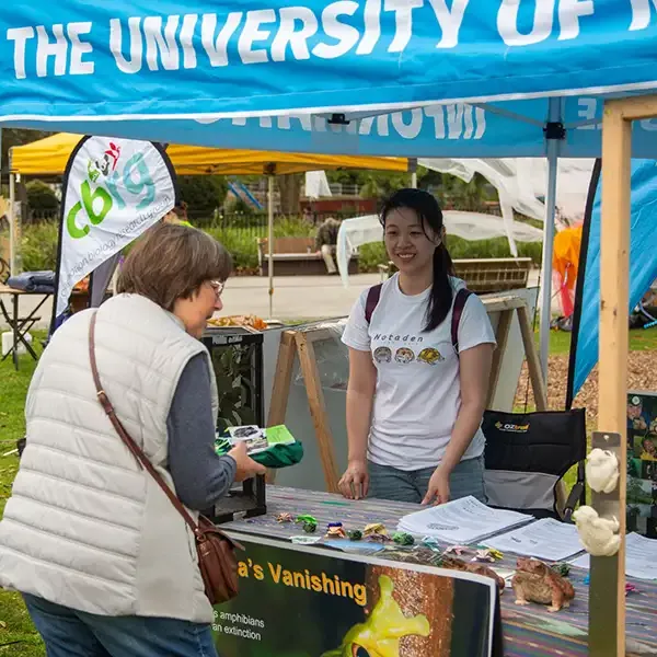 A University representative behind a counter at a University festival stall talking to a festival attendee.