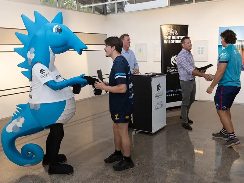 The blue University of Newcastle seahorse mascot and an event speaker interact with athletes during a presentation.