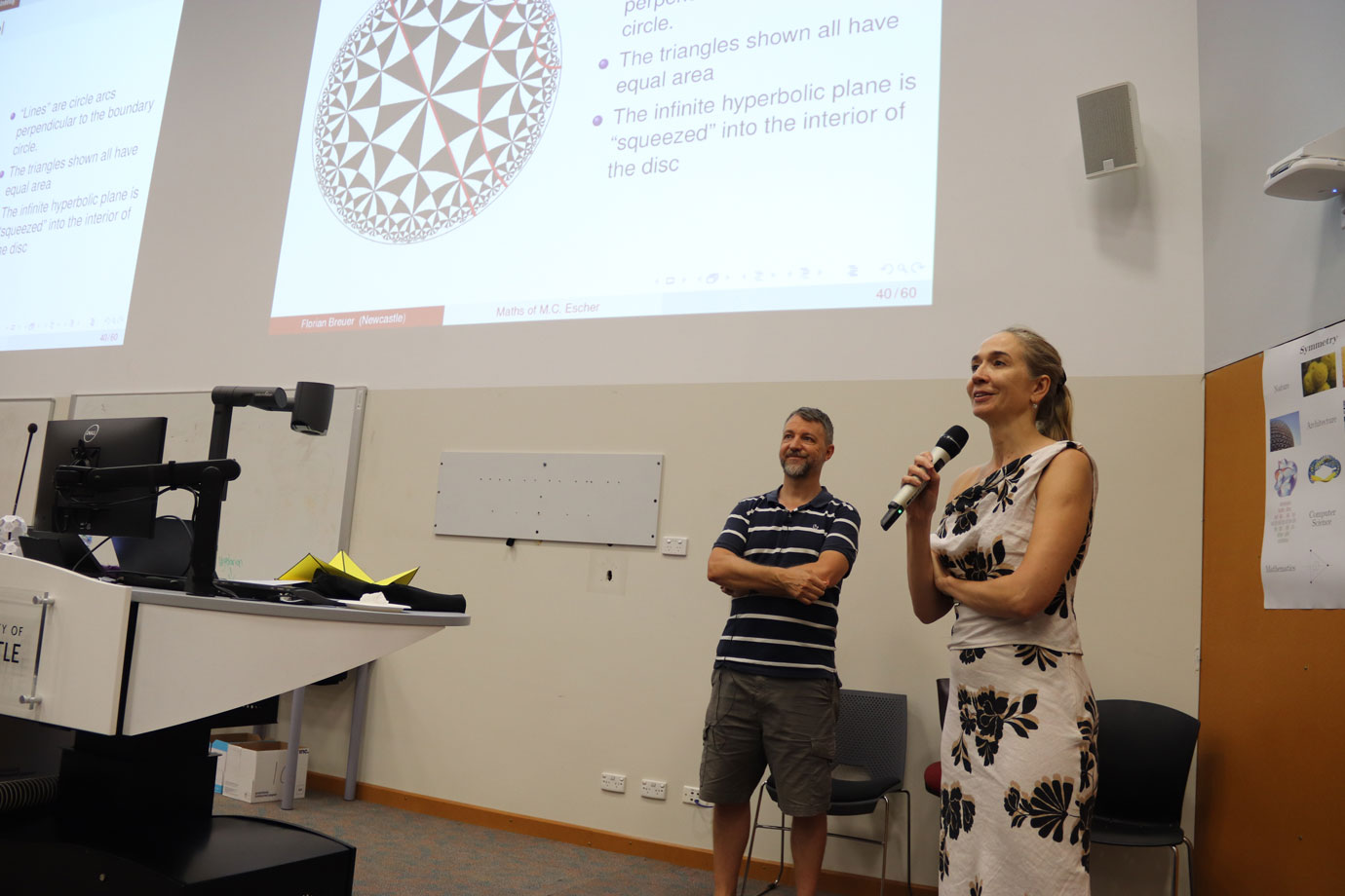 Two Academics smile, looking out into the audience, standing below the presentation projection