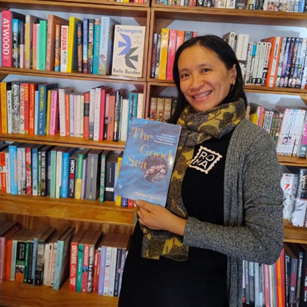A person stands in front of a wall of colorful bookshelves, holding a book titled “The Good Sea” and wearing a patterned scarf and dark outfit.