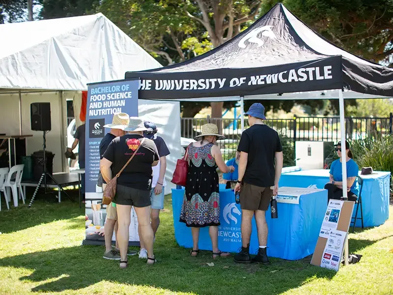 A small crowd of festival attendees standing in front of a University festival stall with a sign in front labelled 'Bachelor of Food Science and Human Nutrition'