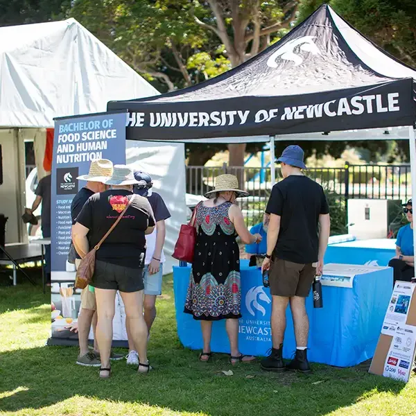 A small crowd of festival attendees standing in front of a University festival stall with a sign in front labelled 'Bachelor of Food Science and Human Nutrition'