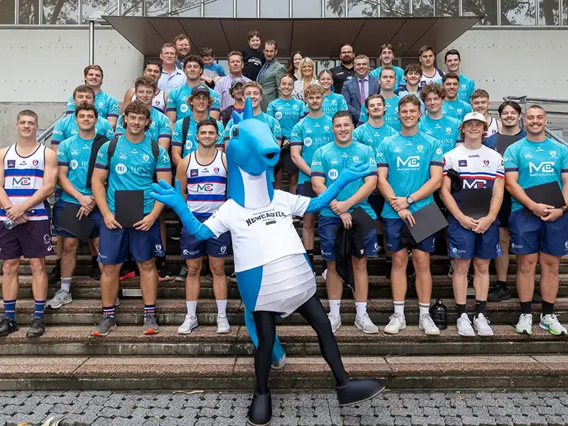 The Hunter Wildfires team poses on outdoor steps with the blue University of Newcastle seahorse mascot striking a pose in front.