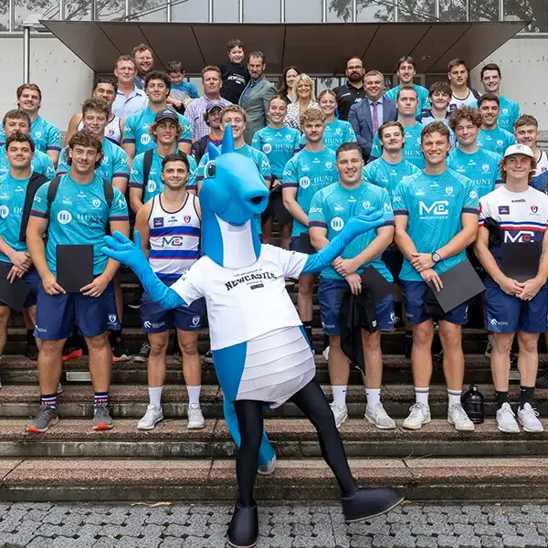 The Hunter Wildfires team poses on outdoor steps with the blue University of Newcastle seahorse mascot striking a pose in front.