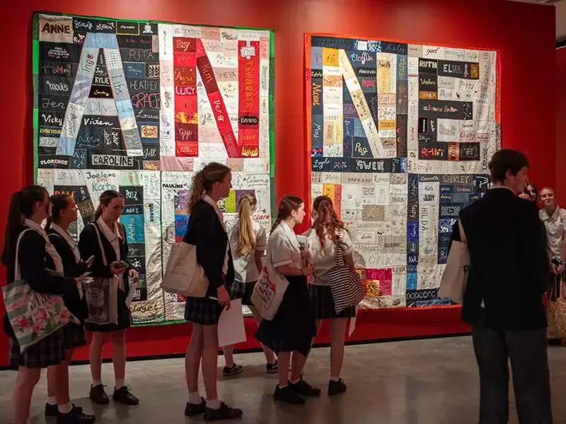 A small crowd of students walking past an artwork on a wall with letters spelling 'Anne' visible in separate panels.