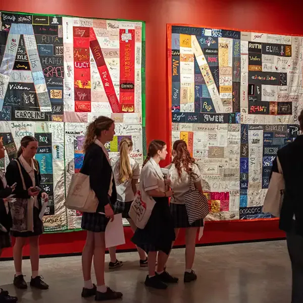 A small crowd of students walking past an artwork on a wall with letters spelling 'Anne' visible in separate panels.