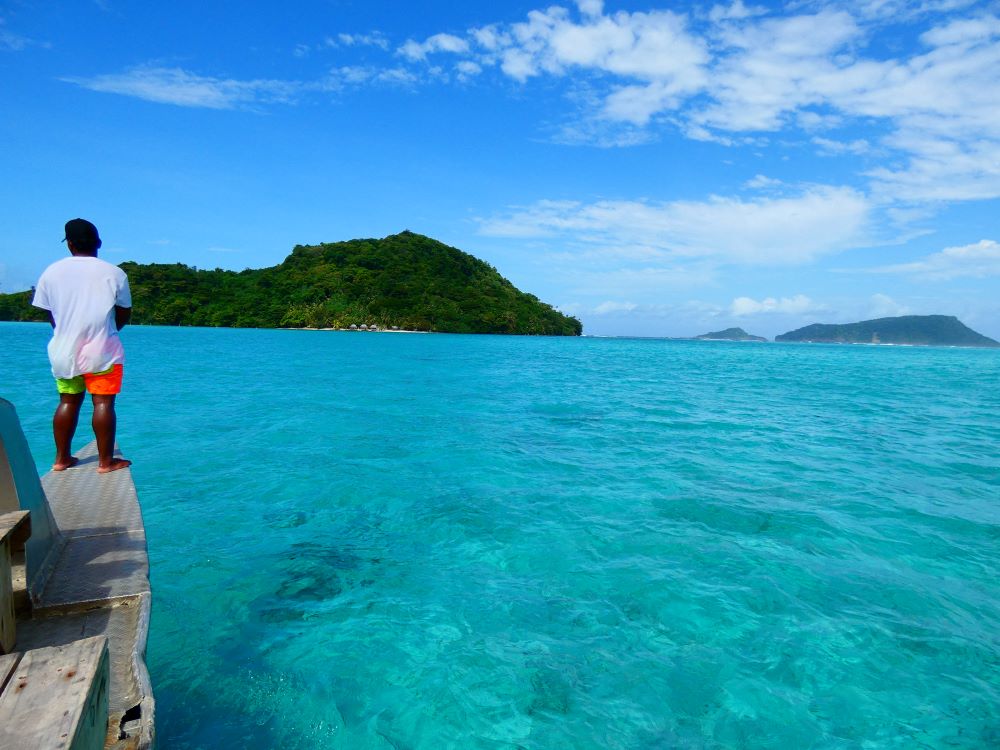 Pristine blue water lagoon with an Island in view. A man is pictured at the end of the boat looking out towards the island. 