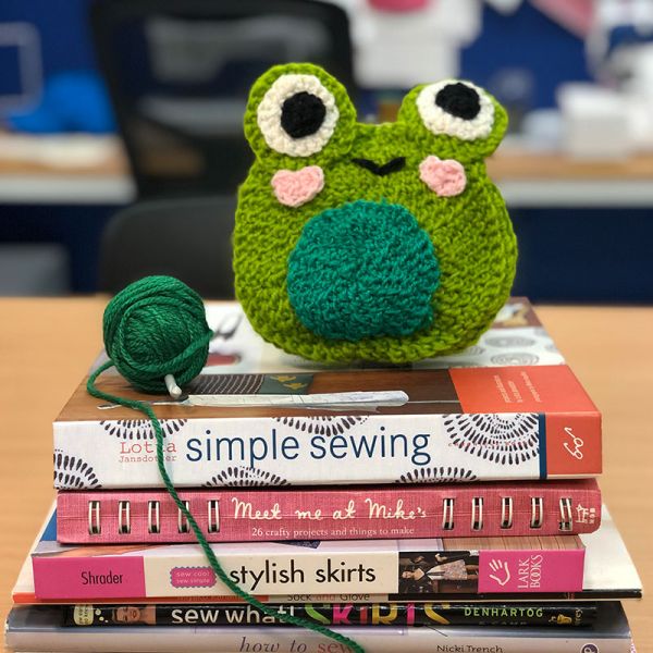 A whimsical crocheted frog sits atop a stack of sewing and crafting books