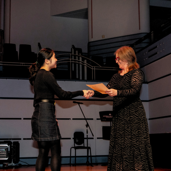 Two individuals stand on a stage as one hands a document to the other during what appears to be an award or presentation ceremony.