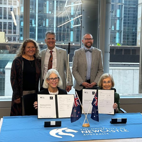 group of leaders smiling to camera, sitting and standing around a table with University of Newcastle blue tablecloth. Partnership to grow Pacific research pipelines and accelerate Pacific led solutions.