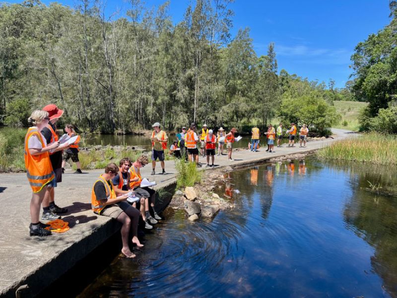 Students sit and stand along the waterside in hi-vis vests with clipboards. 
