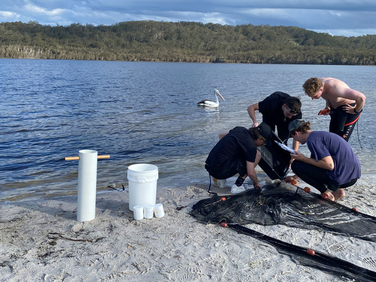 Four students examine equipment on a beach. In the background a pelican floats along the water.