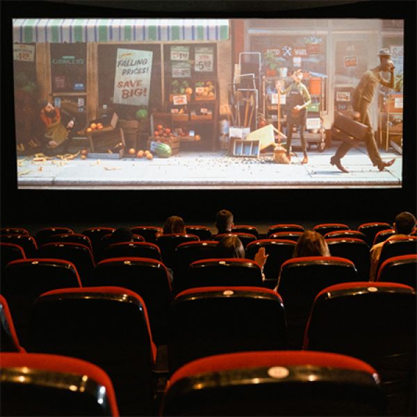 View from the back of a cinema auditorium with red seats showing a projected animated scene of a cluttered grocery storefront where produce is scattered and a person walks past while another stands nearby.
