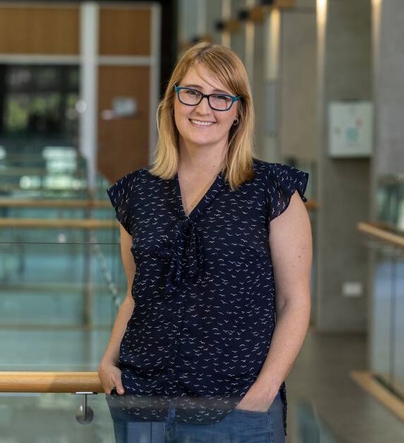 Woman with blonde hair and glasses standing with hands in her pockets and smiling for the camera
